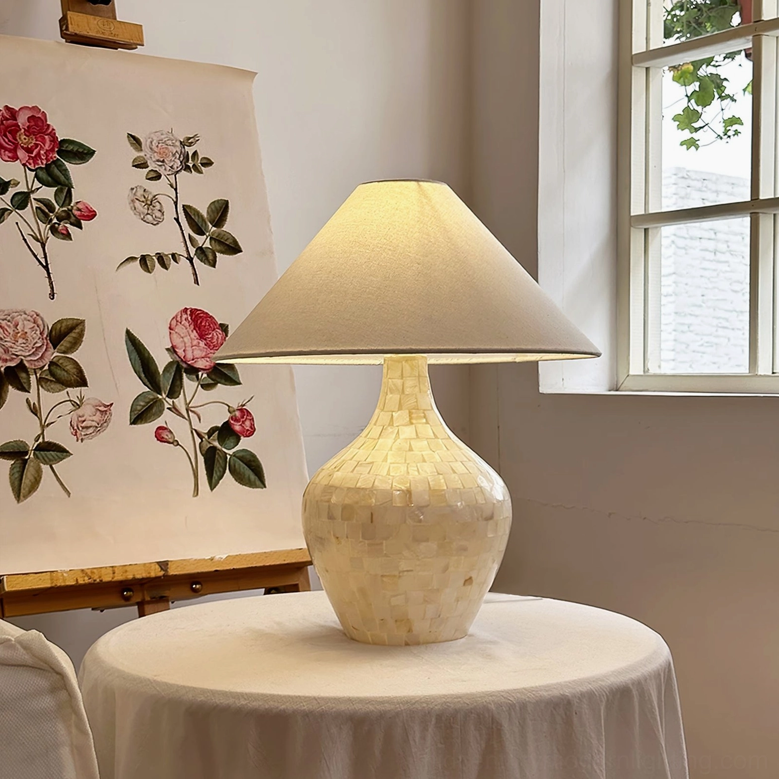 Handcrafted seashell table lamp on round art studio table with floral painting and window light, creative workspace