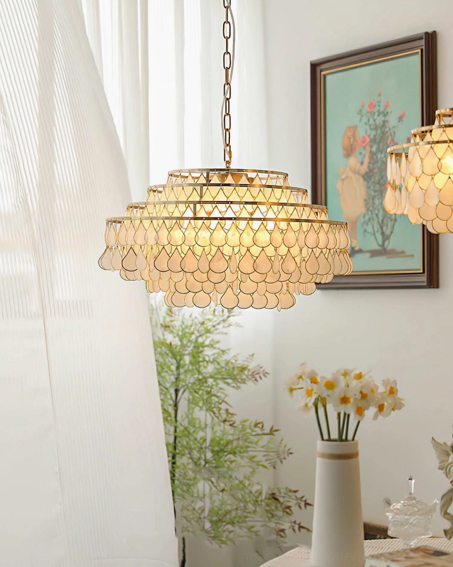 Tiered shell pendant lamp side view behind sheer curtain, indoor space with framed wall art, flowers and green plants, gold suspension chain