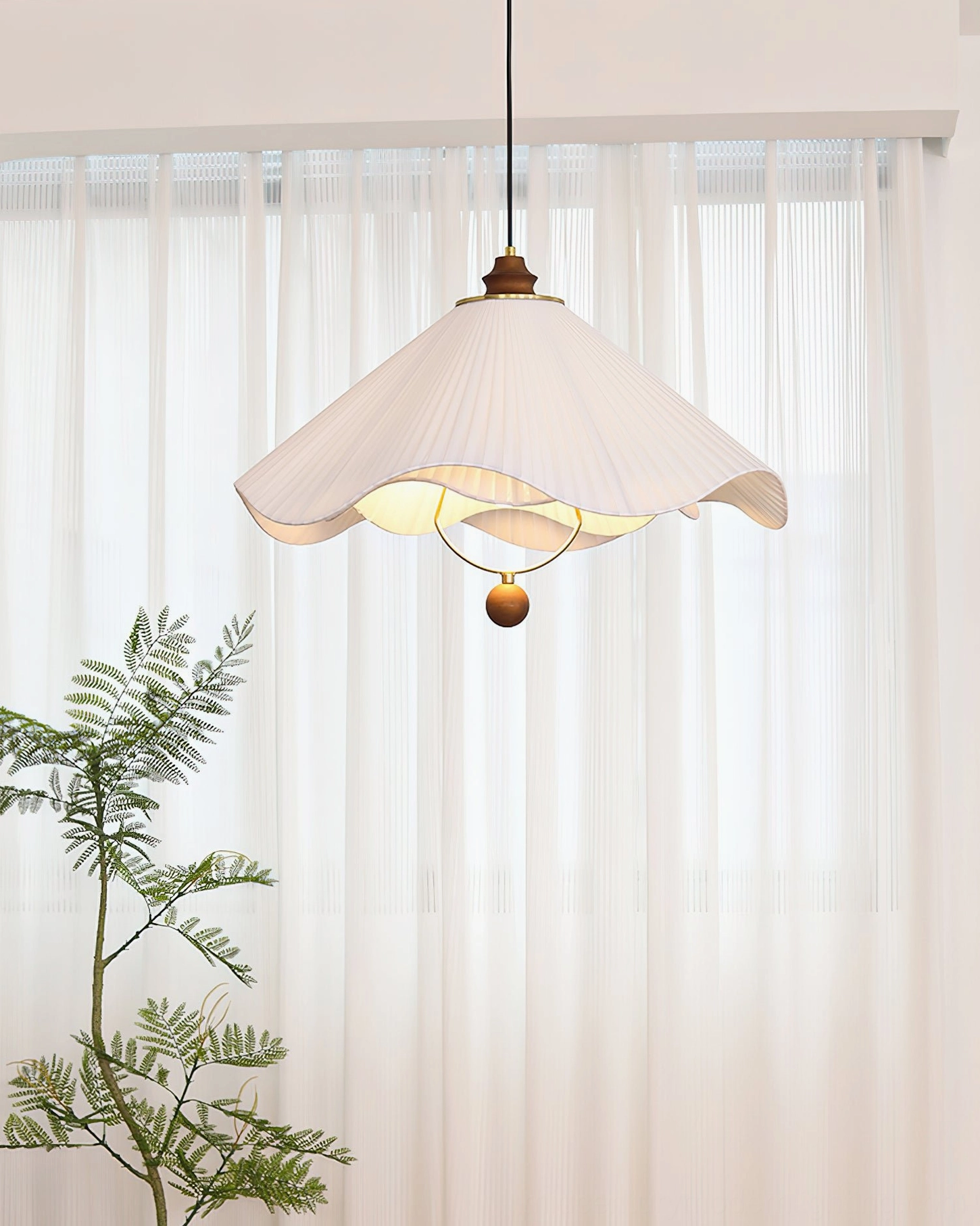 Sloane pleated pendant lamp top-down side view with sheer curtain and potted fern backdrop showing compact size and warm ambient glow