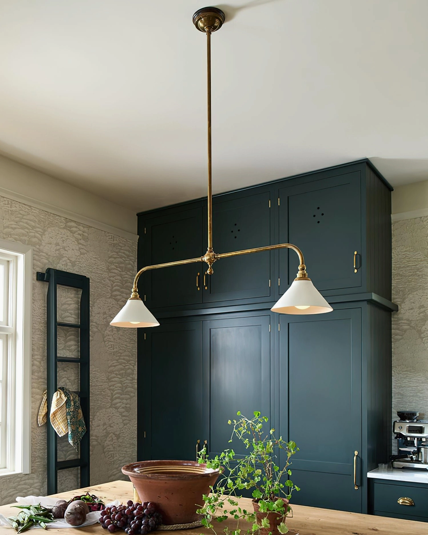 Eye-level view of Theron brass pendant light in farmhouse kitchen with dark green cabinetry, white conical shades providing task lighting over dining surface