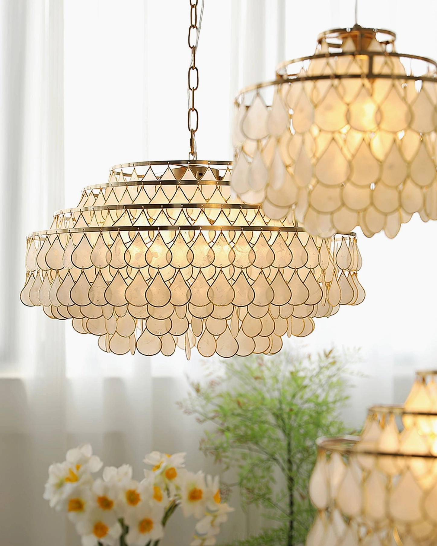 Multiple tiered shell pendant lamps in close-up, indoor setting with sheer curtains, floral and green plant decor, warm glowing vintage lighting