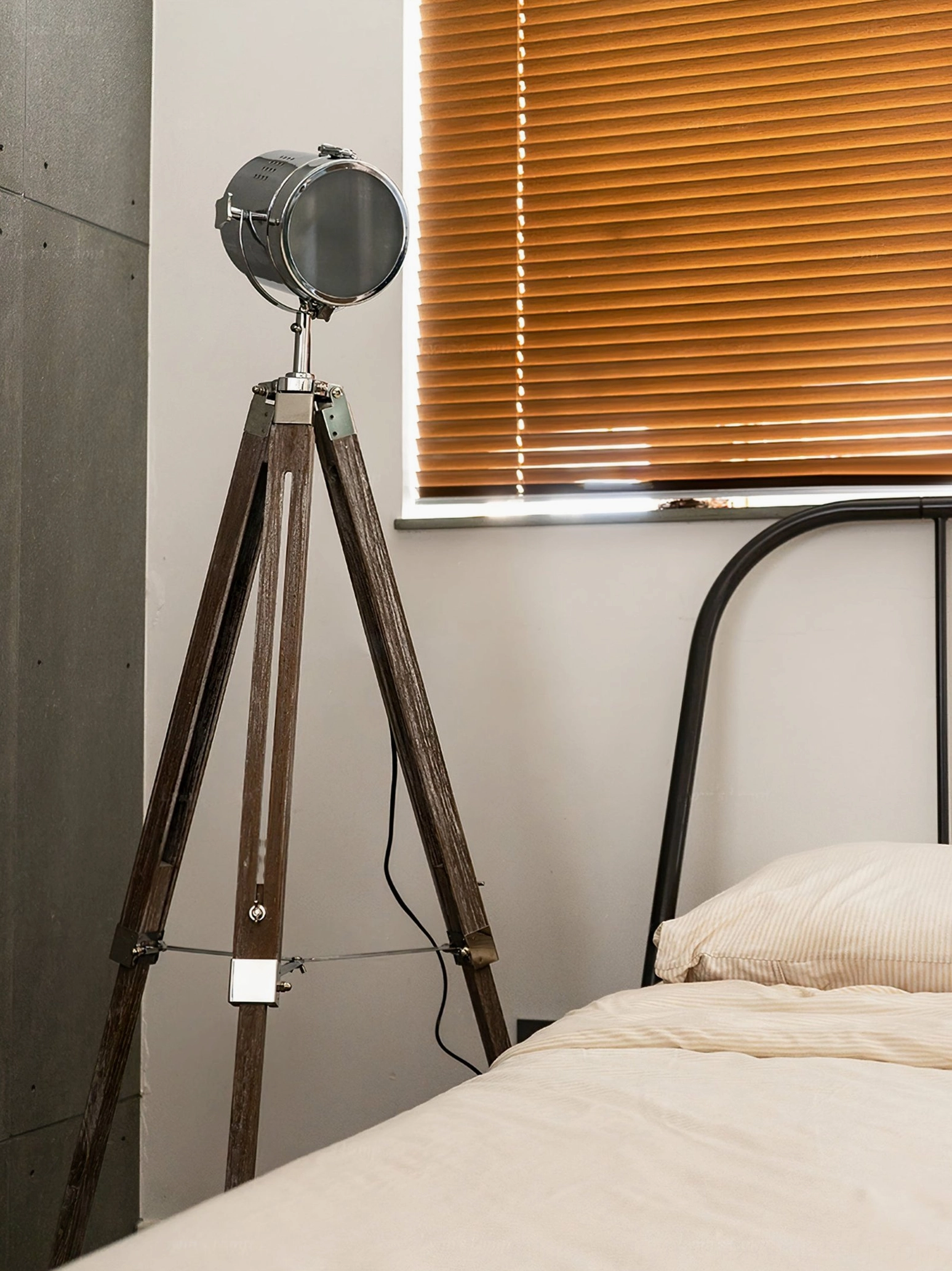 Industrial tripod floor lamp side-rear view in bedroom setting with iron bed and wooden blinds, unlit fabric shade detail