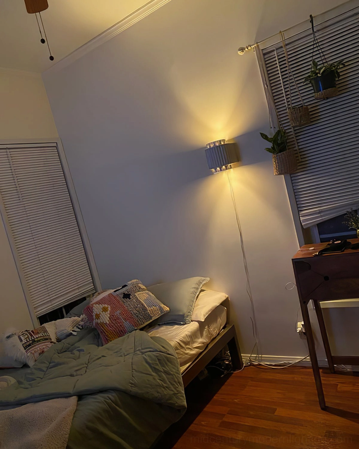 Plug-in Tristan wall lamp providing warm light in cozy bedroom workspace with mixed bedding and hanging indoor plants