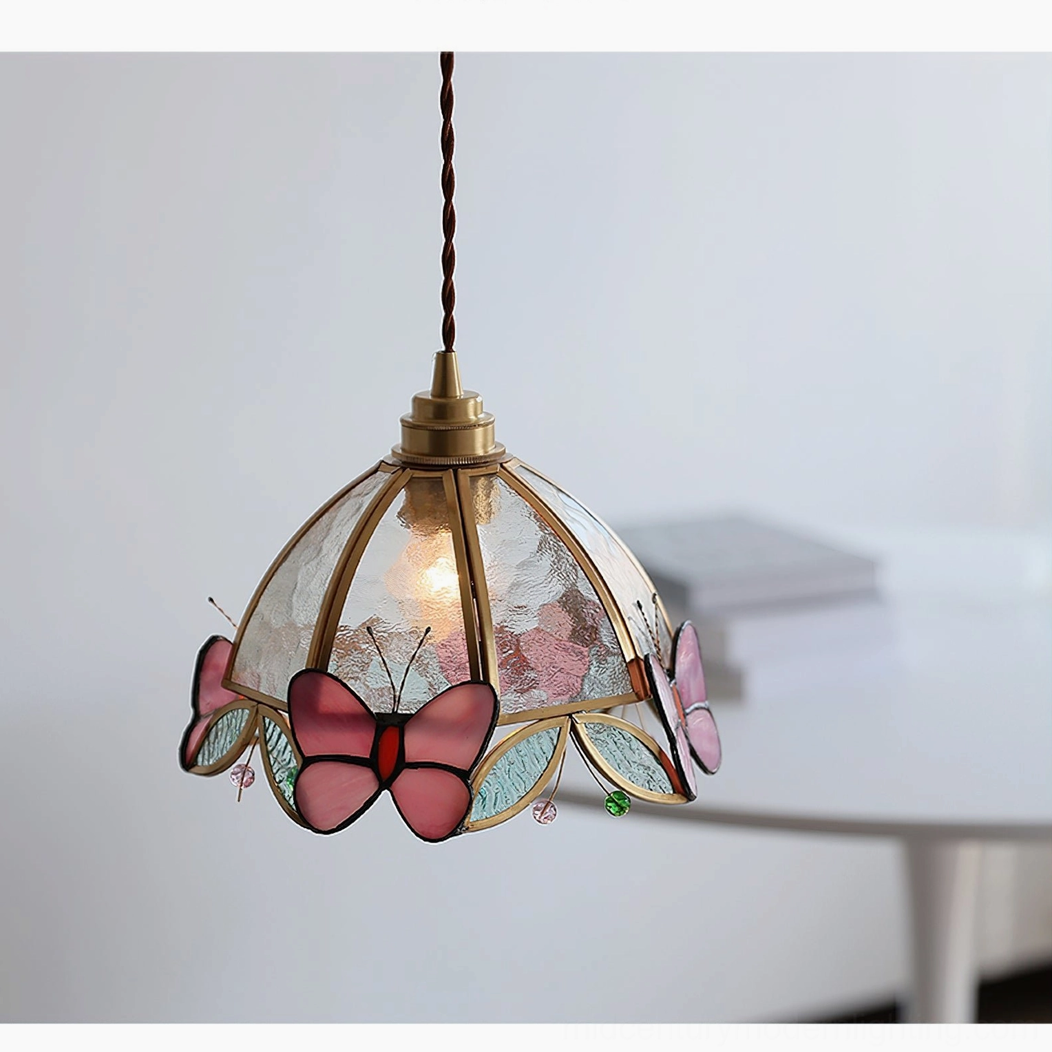 Low-angle front view of Valinte pendant lamp showing large pink butterfly stained glass decor and warm glowing bulb above round white side table
