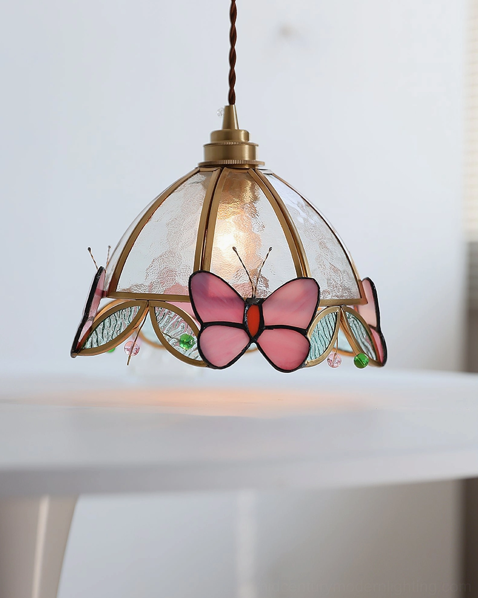 Three-quarter perspective of Valinte butterfly pendant light with twisted brass cord suspended above white side table, vintage Tiffany style design