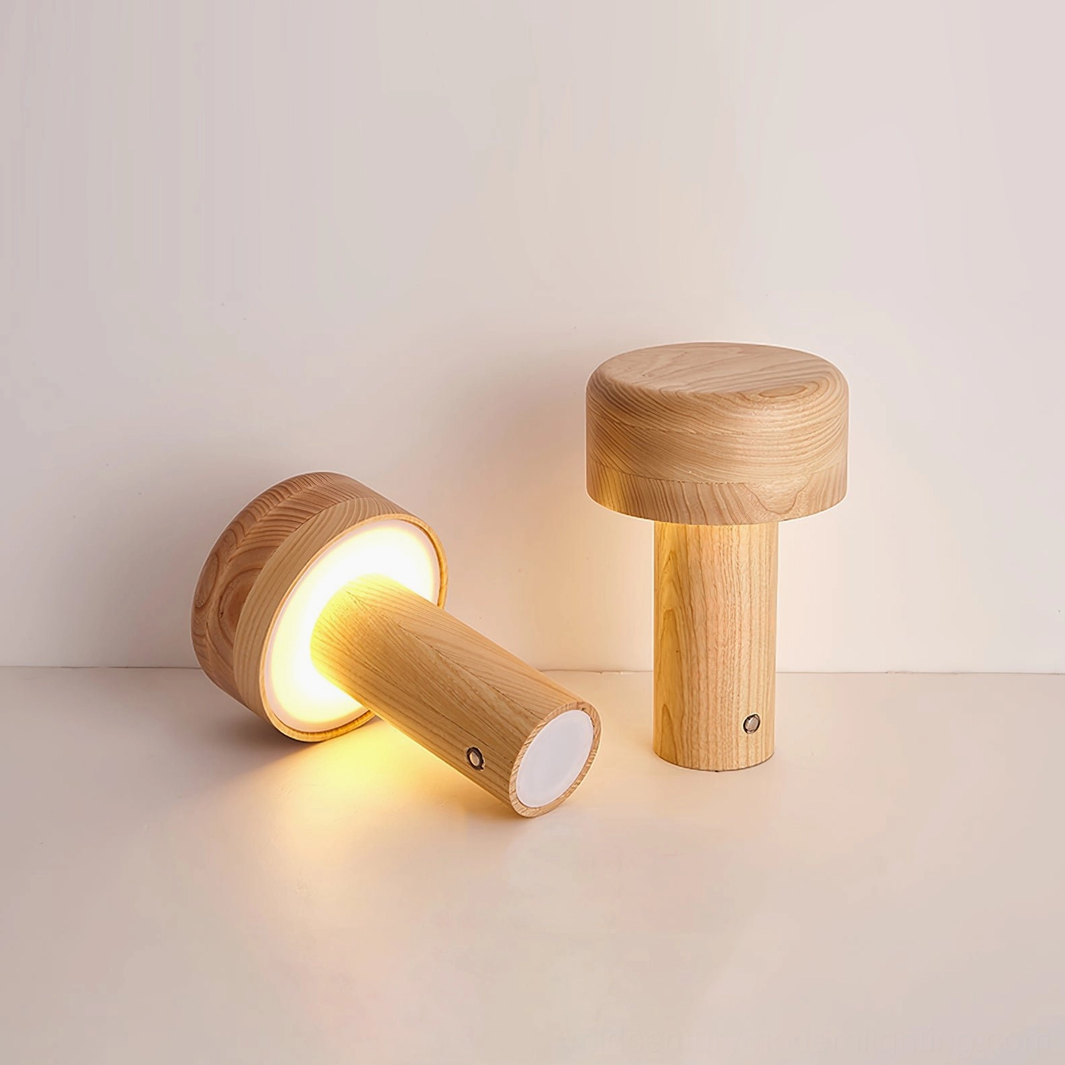 Two wooden cylindrical table lamps on white surface with one lamp lit showing warm glow